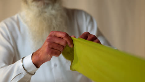 Close-Up-Studio-Shot-Of-Senior-Sikh-Man-With-Beard-Folding-Fabric-For-Turban-Against-Plain-Background-In-Real-Time